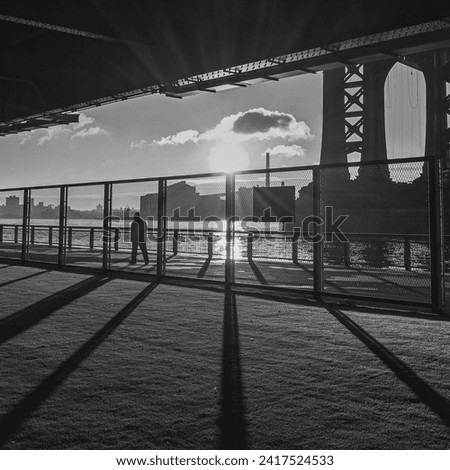 black and white square photo underneath the FDR roadway in new york city at sunrise casting long shadows with a silhouette of a single person walking