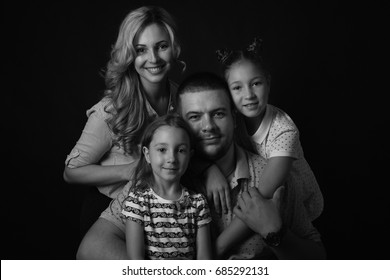 Black And White Stufio Portrait Of Young Family Hugging And Posing On Dark Studio Background