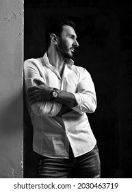 Black And White. Unshaved Man In White Shirt With Rolled Up Sleeves And Jeans Stands With Arms Crossed At Chest, Leaning On Concrete Wall And Looks Aside Over Black Background