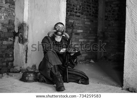 Black and white view of aggressive man with long cloak, machine gun and  mask on his face before fight sits inside old abandoned building 