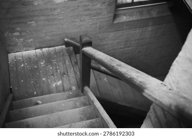 Black and white view down a worn wooden stairway inside an old building, with handrail, brick walls and soft light creating a moody abandoned feel.