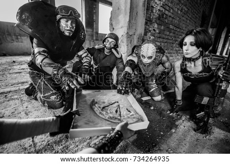 Black and white view of group aggressive men in leather armor, weapon and metal spiked mask on their faces eat pizza before fight inside old abandoned building 