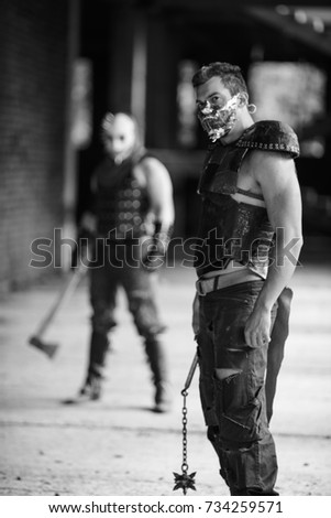 Black and white view of two aggressive men in leather armor, weapon and metal spiked mask on their faces berore fight inside old abandoned building 
