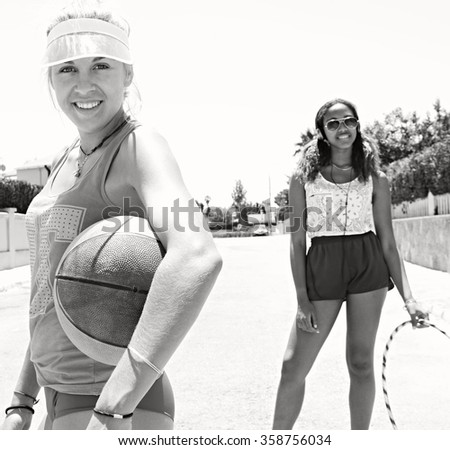 Black and white view of two diverse teenager friends enjoying a holiday together in suburban street, sporty with basketball, hoola hoop and headphones technology. Retro adolescent lifestyle outdoors.