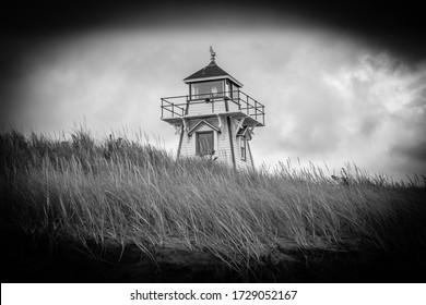 A Black And White Vignette Photo Of A Classic 19th Century Wooden Lighthouse On Top Of Grassy Sand Dunes On The Atlantic Coast Under Storm Clouds.