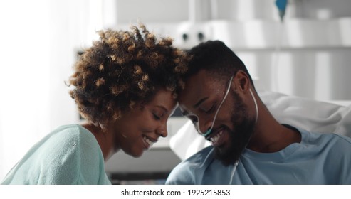 Black Wife Visiting And Talking With Patient Husband In Hospital Bed. Close Up Portrait Of Smiling Young African Woman And Ill Man Cuddling And Chatting In Hospital Ward