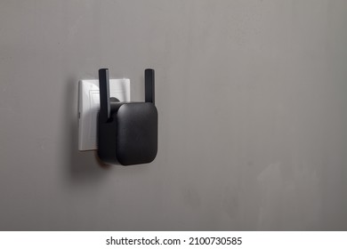 A black Wi-Fi signal amplifier is plugged into an outlet on a gray wall background. Copy Space.