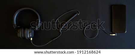 Black wired headphones with ear pads and a smartphone lie on a black background. Analog wire connection using mini-jack - 3.5 mm. Music and creativity. Banner