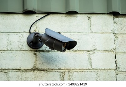 Black wired surveillance anti-theft camera mounted onto cream brick wall near green metal roofing