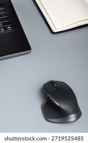 A black wireless computer mouse on a gray desk beside a laptop and an open notebook.