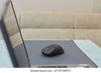 Black wireless computer mouse on a mat next to a laptop, Modern workplace at home or in the office, Side view of a computer mouse and part of a laptop on a table, remote work concept.