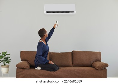 Black woman adjusts temperature settings of her AC at home. Young Afro American lady sitting on comfortable brown sofa uses remote control to turn on white air conditioner on gray wall - Shutterstock ID 2146691889