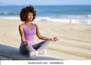 Schwarze Frau, die nach dem Friseur Yoga am Strand mit geschlossenen Augen macht. Junge Weibchen, die Sportkleidung in Lotus tragen Figur mit entfochtenem Hintergrund.