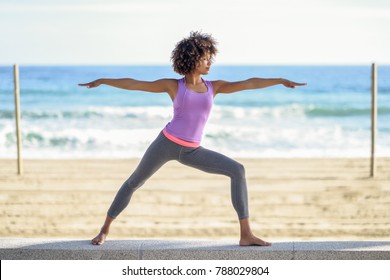 Black woman, afro hairstyle, doing yoga in warrior pose in the beach. Young Female wearing sport clothes with sea at the background