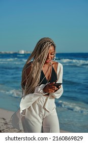 Black Woman With Braids Walking On Beach In Summer With Wide Beach Outfit. Talking On Cell Phone And Happy Expression. Walking Barefoot. Vertical Photo