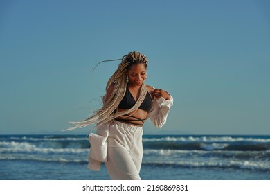 Black Woman With Braids Walking On The Beach In Summer With A Wide Beach Outfit. Smiling With A Happy Expression. Horizontal Photo