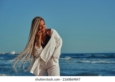 Black Woman With Braids Walking On The Beach In Summer With A Wide Beach Outfit. Talking On The Cell Phone And With A Happy Expression. Walking Barefoot. Photo Vertical