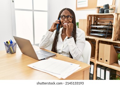 Black Woman With Braids Working At The Office Speaking On The Phone Looking Stressed And Nervous With Hands On Mouth Biting Nails. Anxiety Problem. 