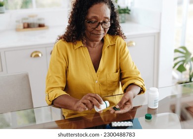 Black woman with a chronic health condition sits at her kitchen table taking her medication. Senior woman adhering to her prescribed treatment in order to efficiently manage her personal health.