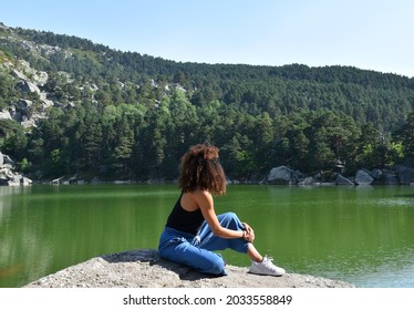 Black woman with curly afro hair sitting on top of a rock. Observing the Black Lagoon and the pine forest in Soria, Spain.