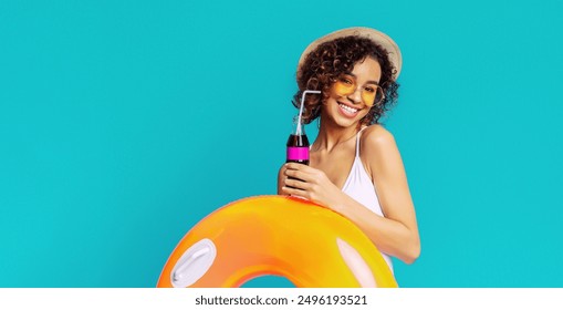 Black woman with curly hair smiles brightly as she poses in front of a blue backdrop. She is holding a soda with a straw and an inflatable ring, suggesting a day of fun and relaxation in the sun. - Powered by Shutterstock