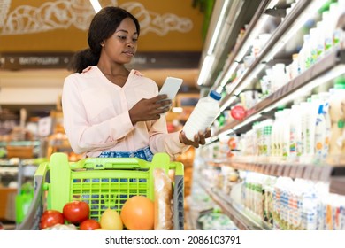 Black Woman Customer With Smartphone Scanning Bottle Of Milk During Shopping Buying Groceries Via Mobile Application Standing With Cart In Supermarket. Modern Shopping Concept