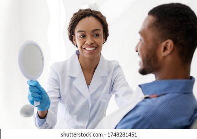 Black Woman Dentist Doctor Showing Young Man Patient Mirror To Check His New Smile, Dental Clinic Interior