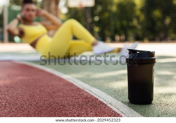 Black\
woman doing abs exercises crunches with arms behind head on floor\
yoga mat, working on six-pack abs in blurred background, selective\
focus on black shaker on basketball field\
ground