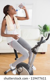 Black Woman Doing Exercise Bike While Drinking In A Living Room