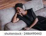 Black woman in a black dress and headwrap lounges on a gray couch, resting her head on her hand and smiling gently