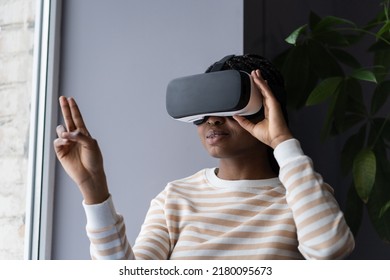 Black Woman Enjoying Virtual Reality Shopping Experience While Sitting By Window At Home, African American Female Wearing VR Helmet Touching Air With Finger, Testing Modern Hi-tech Future Gadget