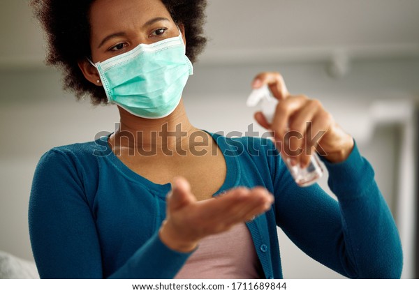 Black woman with face mask using hand\
sanitizer at home during virus pandemic.\
