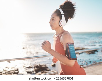 Black woman, fitness and running with headphones and cellphone on arm at the beach in Cape Town. Sporty African American female runner by the ocean coast having a run for cardio training workout - Shutterstock ID 2256589801