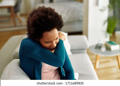 Black woman with flu virus sneezing into elbow in the living room.  - Powered by Shutterstock