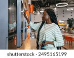 Black Woman with glasses and striped shirt using a touch screen kiosk in a modern shopping mall. Girl chooses burger on touch menu of fast food restaurant. 