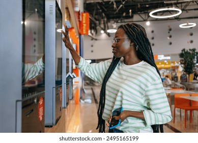 Black Woman with glasses and striped shirt using a touch screen kiosk in a modern shopping mall. Girl chooses burger on touch menu of fast food restaurant. 