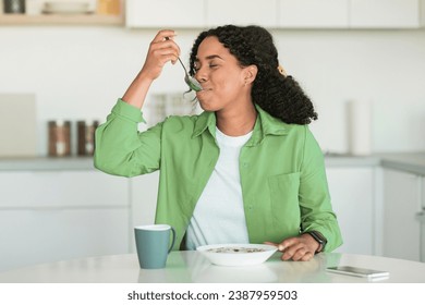 Black woman in green shirt starts her day with tasty breakfast in modern kitchen, sitting at dining table and savoring healthy cereals flakes and coffee, enjoys balanced meal and nutritious living