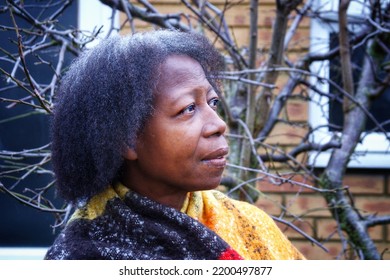 A Black Woman With Greying Hair Wearing A Colourful Scarf In Focus Against The Backdrop Of A Garden Shed With Brick Wall And A Leafless Tree In Soft Focus