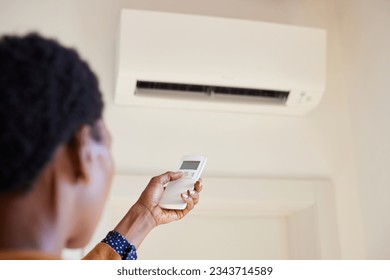 Black woman hand adjusting AC temperature with remote control to wall type air conditioning home cooling system. Hand of african woman holding remote controller directed towards the air conditioner.