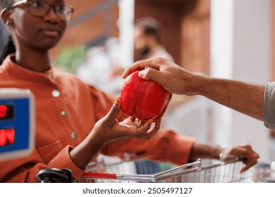 A black woman hands a fresh red bell pepper to a shopkeeper to weigh. African American customer checks the weight of locally grown fruits and vegetables at the cashier's desk. - Powered by Shutterstock