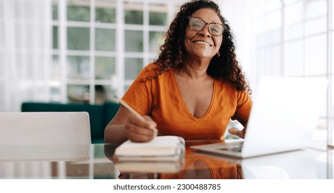 Black woman happily planning her retirement from the comfort of her own home. Mature woman sitting at a table with her laptop, writing her goals and dreams for the future in a journal. - Powered by Shutterstock