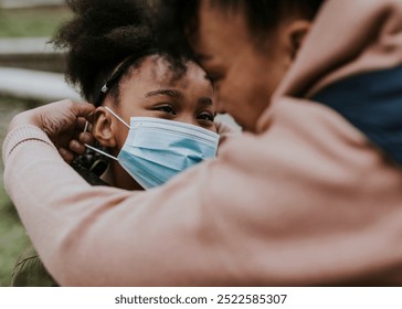 A Black woman helps a Black happy girl adjust her face mask. The woman and girl child share a tender happy moment. The happy girl kid looks up at the Black woman while adjusting the mask. - Powered by Shutterstock