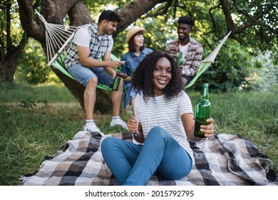 Black Woman Holding Bottle Of Beer And Grilled Sausages In Skewer While Resting At Green Garden. Three Multiracial Friends Chatting On Background.