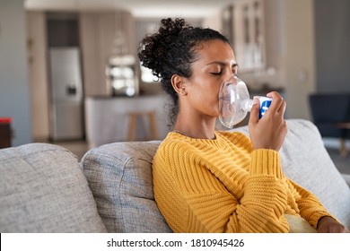 Black woman holding a mask nebulizer inhaling fumes medication into lungs. African sick lady inhaling through inhaler mask at home. Self treatment of the respiratory tract using inhalation nebulizer.