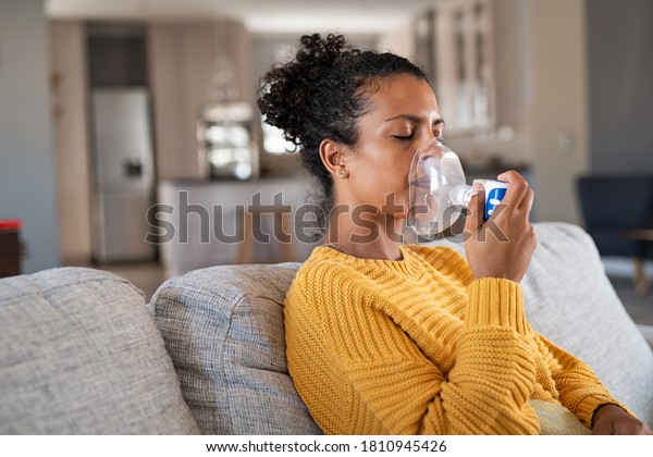 Black woman holding a mask nebulizer inhaling\
fumes medication into lungs. African sick lady inhaling through\
inhaler mask at home. Self treatment of the respiratory tract using\
inhalation nebulizer.