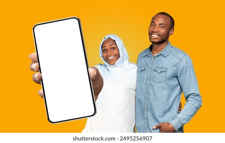 Black woman in a light blue hijab smiles and points to a blank phone screen while standing next to a man in a denim shirt. Both are looking directly at the camera. - Powered by Shutterstock