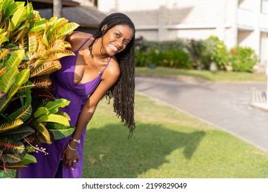 Black Woman With Long Natural Hair Smiling Happily Peeking Out From Behind A Croton Plant