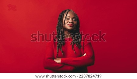 Black woman looking at the camera while standing against a red background with her arms crossed. Mature woman with dreadlocks embracing her natural hair with confidence.