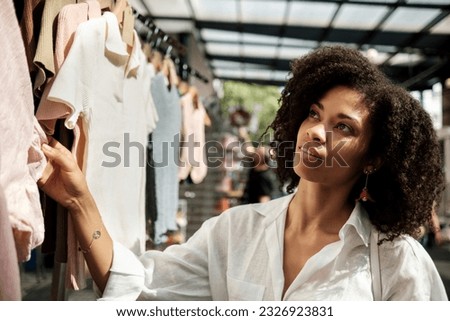 Black woman looking outfits in street market. There are many items hanging on hangers. She is wearing a fresh white shirt and some jewelry. Traditional commerce.
