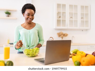 Black Woman Making Salad At Laptop Computer Learning To Cook Online In Modern Kitchen At Home. Happy Housewife Preparing Healthy Dinner And Browsing Recipes. Selective Focus